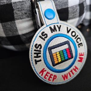 Puede incluir: Llavero bordado con el texto "THIS IS MY VOICE KEEP WITH ME" en negro y rojo. El dise&ntilde;o incluye un gr&aacute;fico colorido y un bot&oacute;n azul. El llavero est&aacute; sujeto a un clip plateado.