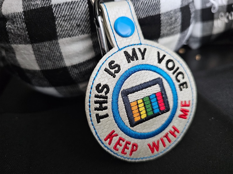 Puede incluir: Llavero bordado con el texto "THIS IS MY VOICE KEEP WITH ME" en negro y rojo. El dise&ntilde;o incluye un gr&aacute;fico colorido y un bot&oacute;n azul. El llavero est&aacute; sujeto a un clip plateado.
