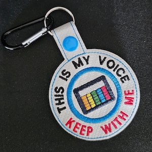 May include: Embroidered keychain with the text "THIS IS MY VOICE KEEP WITH ME". The round, white keychain features a blue circle with a colorful graphic and a black carabiner clip.