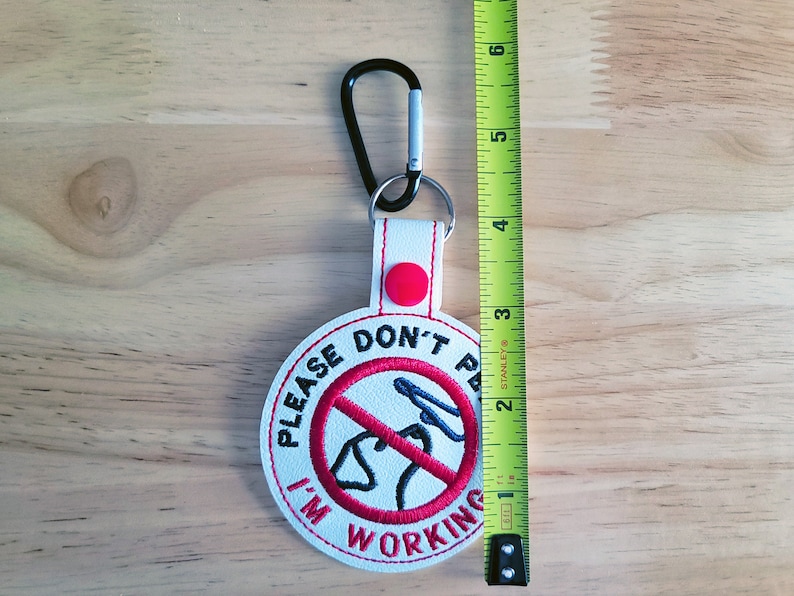 May include: White embroidered keychain with the text "PLEASE DON'T PET I'M WORKING" in a red circle with a dog silhouette. The keychain has a black carabiner clip and a red button. The keychain is approximately 3 inches long.