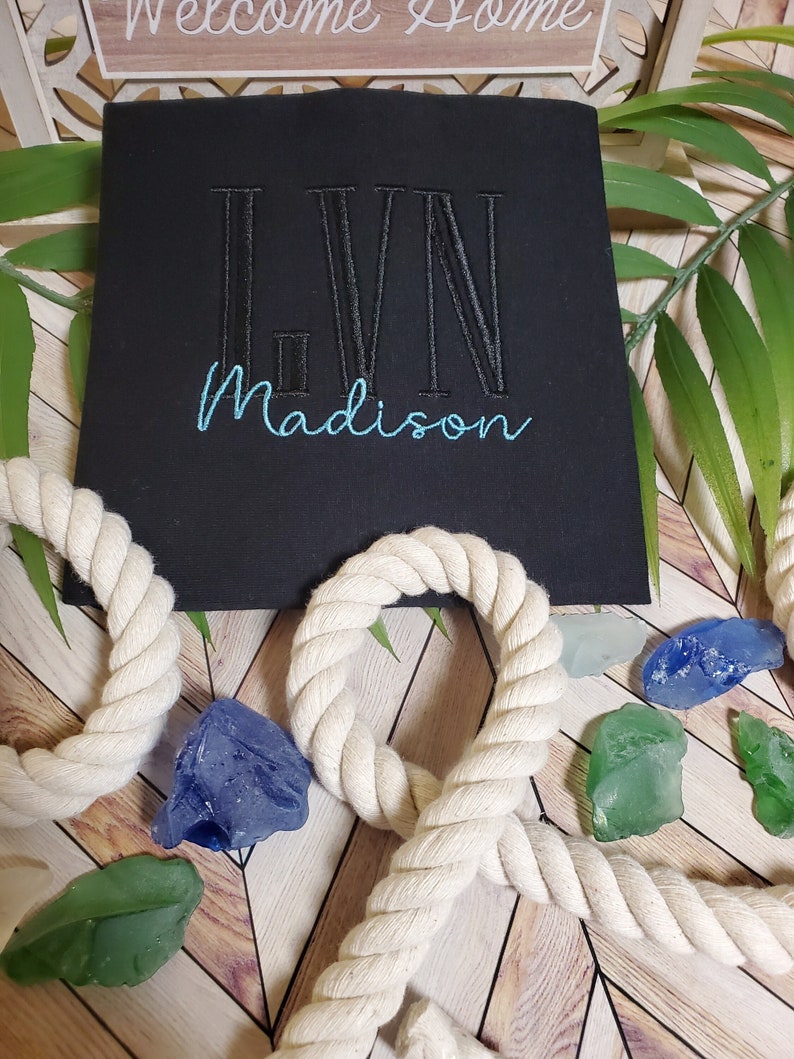 May include: A black t-shirt featuring the embroidered monogram LVN in black thread and the name Madison in light blue cursive script. The shirt is displayed with decorative white rope and scattered blue and green glass stones.
