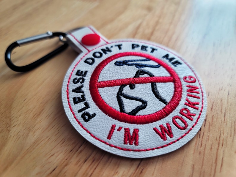 May include: A round, white embroidered patch with the text "PLEASE DON'T PET ME I'M WORKING" in red. The patch features a red circle with a crossed-out hand and dog illustration. It has a black carabiner clip and a red snap.