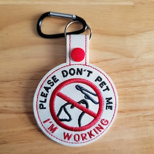 May include: White and red embroidered dog tag with the text "PLEASE DON'T PET ME I'M WORKING". The tag features a black carabiner clip and a red button. The design includes a red circle with a dog silhouette crossed out.