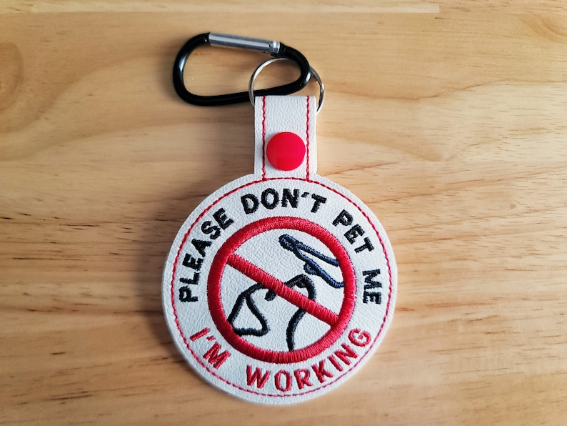 May include: White and red embroidered dog tag with the text "PLEASE DON'T PET ME I'M WORKING". The tag features a black carabiner clip and a red button. The design includes a red circle with a dog silhouette crossed out.