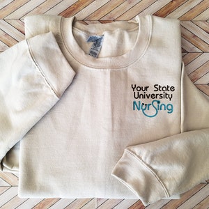 May include: A beige long-sleeve crewneck sweatshirt featuring the embroidered text Your State University in black and the word Nursing in blue, with the letter u in Nursing stylized to resemble a stethoscope.