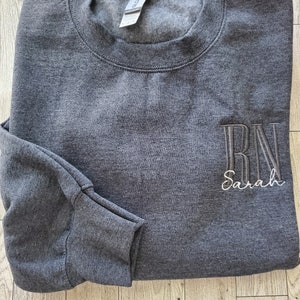 May include: A dark charcoal gray crewneck sweatshirt featuring long sleeves and a ribbed collar. The front right chest displays embroidered text in a vertical block font reading RN, with the name Sarah embroidered below in a white script font.