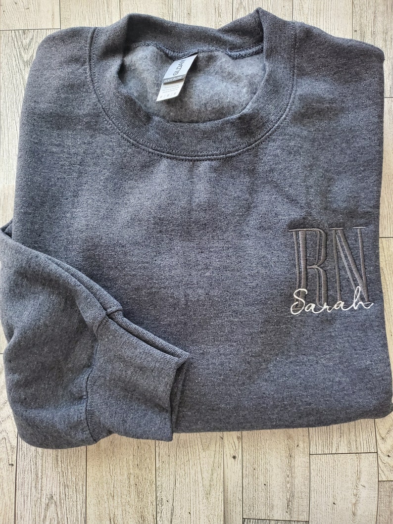 May include: A dark charcoal gray crewneck sweatshirt featuring long sleeves and a ribbed collar. The front right chest displays embroidered text in a vertical block font reading RN, with the name Sarah embroidered below in a white script font.