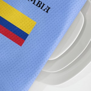 Puede incluir: Una servilleta azul claro con la palabra "COLOMBIA" impresa en negro y la bandera colombiana en amarillo, azul y rojo. La servilleta cubre parcialmente una pila de platos blancos.