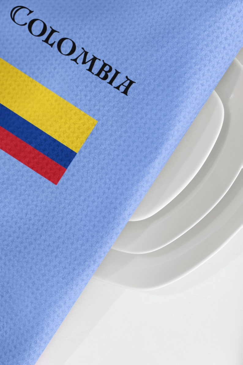 Puede incluir: Una servilleta azul claro con la palabra "COLOMBIA" impresa en negro y la bandera colombiana en amarillo, azul y rojo. La servilleta cubre parcialmente una pila de platos blancos.