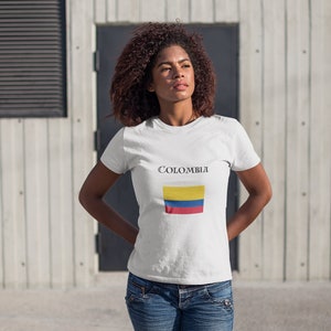 Puede incluir: Camiseta blanca con la palabra "COLOMBIA" impresa sobre un dise&ntilde;o rectangular de la bandera colombiana. La bandera presenta franjas horizontales amarillas, azules y rojas. La camiseta se lleva con vaqueros azules.