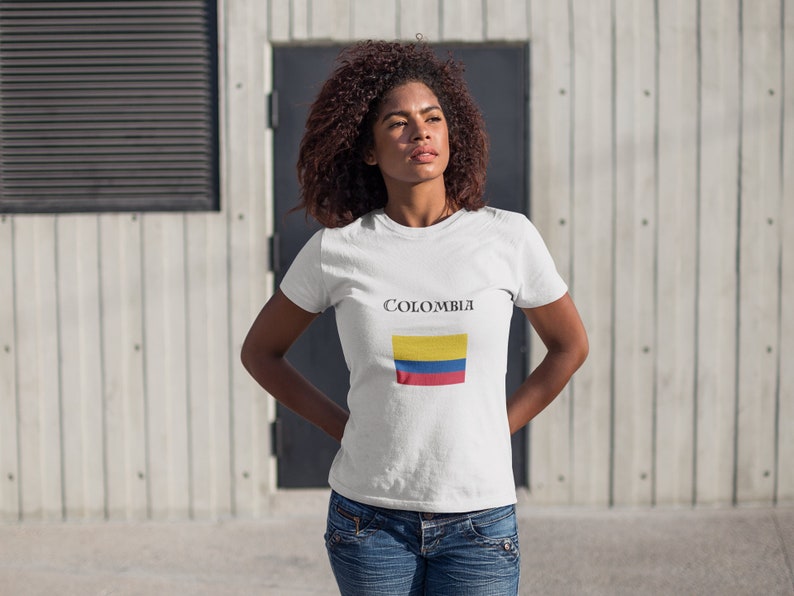 Puede incluir: Camiseta blanca con la palabra "COLOMBIA" impresa sobre un dise&ntilde;o rectangular de la bandera colombiana. La bandera presenta franjas horizontales amarillas, azules y rojas. La camiseta se lleva con vaqueros azules.