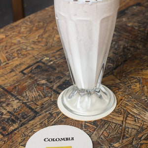 Puede incluir: Un batido en un vaso alto, cubierto con crema batida y chispas de chocolate. Un posavasos con la palabra "COLOMBIA" y la bandera colombiana est&aacute; sobre una mesa de madera. El batido es de color marr&oacute;n claro.