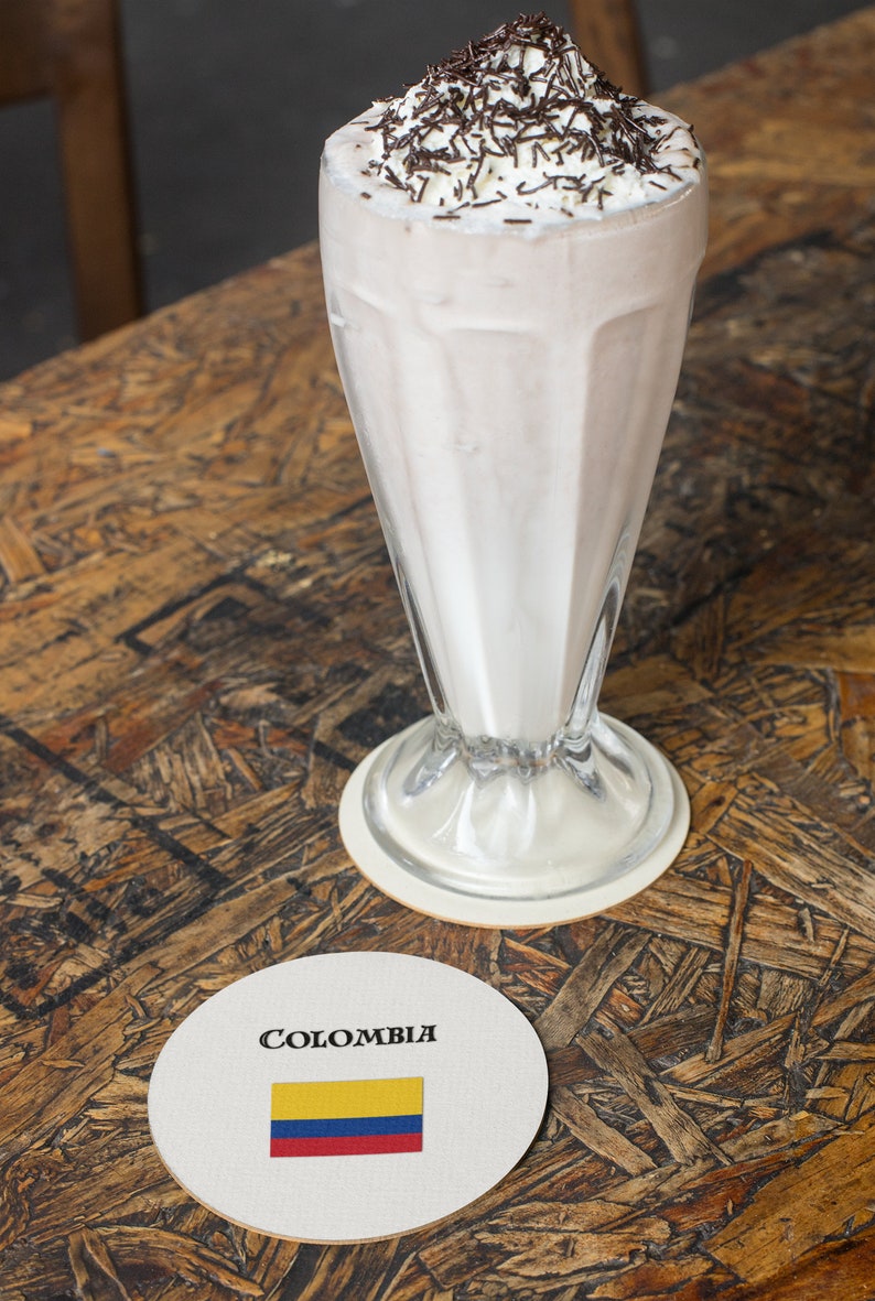 Puede incluir: Un batido en un vaso alto, cubierto con crema batida y chispas de chocolate. Un posavasos con la palabra "COLOMBIA" y la bandera colombiana est&aacute; sobre una mesa de madera. El batido es de color marr&oacute;n claro.