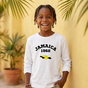 Puede incluir: Una camiseta blanca de manga larga con el texto negro JAMAICA 1962 arqueado sobre un gráfico de la bandera de Jamaica con la forma de la isla. La prenda cuenta con un cuello redondo y mangas lisas para un estilo sencillo y limpio.