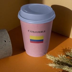 Puede incluir: Una taza de caf&eacute; de papel rosa con tapa azul. La taza lleva la inscripci&oacute;n "Colombia" con la bandera colombiana. La taza est&aacute; sobre una superficie naranja con un bloque de hormig&oacute;n y espigas de trigo.