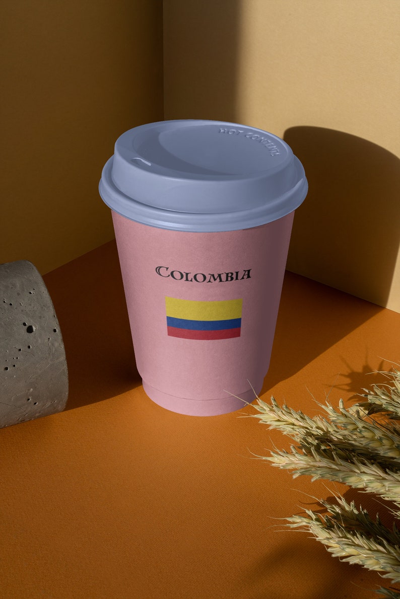 Puede incluir: Una taza de caf&eacute; de papel rosa con tapa azul. La taza lleva la inscripci&oacute;n "Colombia" con la bandera colombiana. La taza est&aacute; sobre una superficie naranja con un bloque de hormig&oacute;n y espigas de trigo.