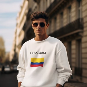 Puede incluir: Sudadera blanca con la palabra "COLOMBIA" sobre un dise&ntilde;o de bandera rectangular en amarillo, azul y rojo. La persona lleva gafas de sol de aviador y pantalones blancos. El fondo muestra una calle de la ciudad.
