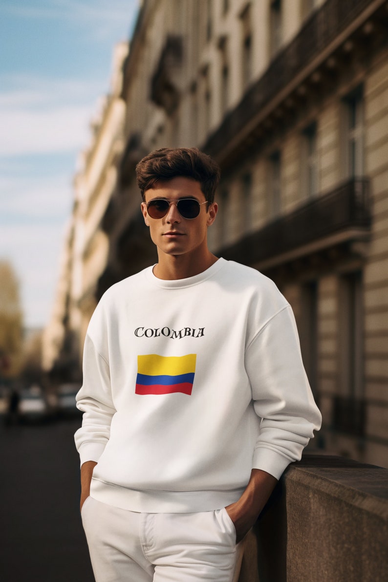 Puede incluir: Sudadera blanca con la palabra "COLOMBIA" sobre un dise&ntilde;o de bandera rectangular en amarillo, azul y rojo. La persona lleva gafas de sol de aviador y pantalones blancos. El fondo muestra una calle de la ciudad.