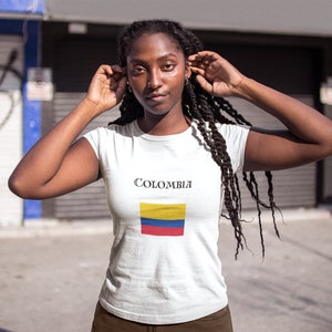 Puede incluir: Camiseta blanca con la palabra "Colombia" y la bandera colombiana. La bandera tiene franjas horizontales amarillas, azules y rojas. La camiseta la lleva una persona con cabello oscuro.