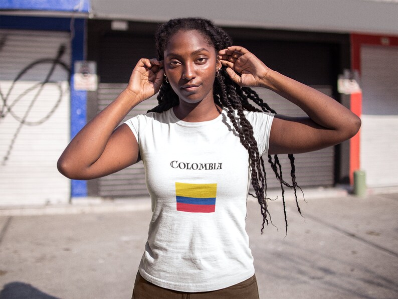 Puede incluir: Camiseta blanca con la palabra "Colombia" y la bandera colombiana. La bandera tiene franjas horizontales amarillas, azules y rojas. La camiseta la lleva una persona con cabello oscuro.