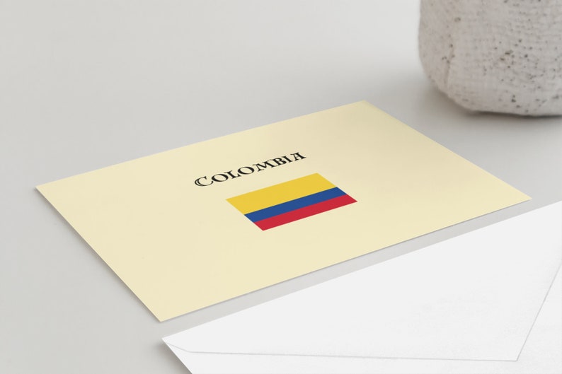 Puede incluir: Una tarjeta color crema con la palabra "COLOMBIA" en escritura negra sobre una bandera rectangular amarilla, azul y roja. Un sobre blanco est&aacute; en primer plano.