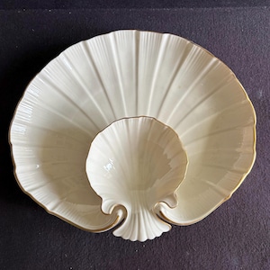 May include: A cream-colored ceramic serving dish shaped like a large scallop shell with a smaller, integrated shell-shaped bowl in the center. The edges of both shell forms are finished with a thin, metallic gold trim, highlighting the radial ridges.