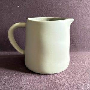 Puede incluir: Una jarra de cerámica verde salvia con un asa curva y un pico vertedor. La jarra tiene un acabado liso y mate y un cuerpo redondeado. Este artículo de cocina es adecuado para servir bebidas.
