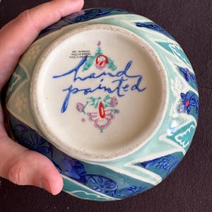 K&ouml;nnte beinhalten: Eine handbemalte Keramikschale mit blauem, t&uuml;rkisem und wei&szlig;em Blumendesign. Die Unterseite zeigt die Worte "hand painted" in blauer Schrift und ein Blumenemblem. Der Rand der Schale hat ein sich wiederholendes Muster aus blauen und t&uuml;rkisen Formen.