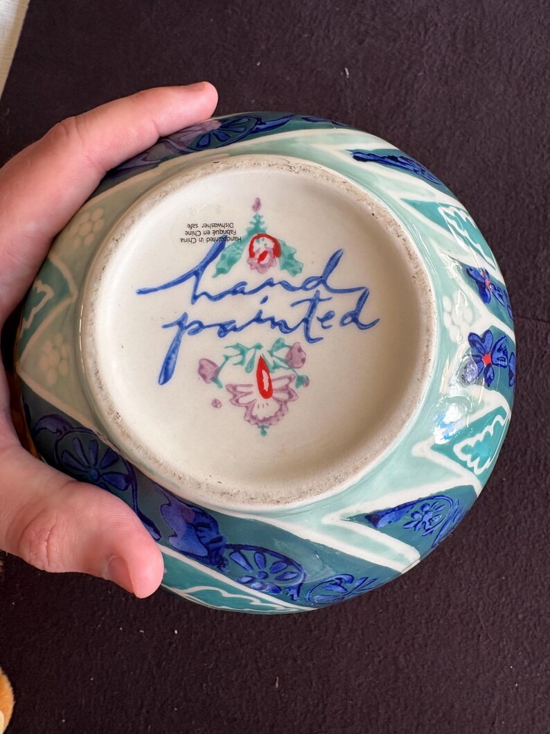K&ouml;nnte beinhalten: Eine handbemalte Keramikschale mit blauem, t&uuml;rkisem und wei&szlig;em Blumendesign. Die Unterseite zeigt die Worte "hand painted" in blauer Schrift und ein Blumenemblem. Der Rand der Schale hat ein sich wiederholendes Muster aus blauen und t&uuml;rkisen Formen.