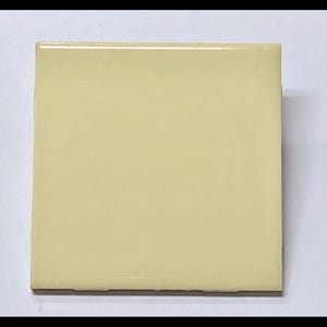 May include: A square, pale yellow tile with a glossy finish. The tile is a solid color and appears to be made of ceramic or a similar material. The edges are slightly rounded.