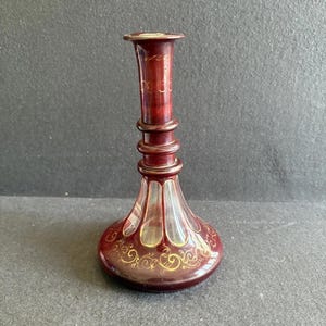 Könnte beinhalten: Eine rubinrote Glasvase mit ausgestelltem Boden und schlankem Hals. Die Vase hat goldene Filigranmuster und Akzente. Die Vase ist etwa 20 cm hoch, mit einem bauchigen Boden und einer schmalen Öffnung.