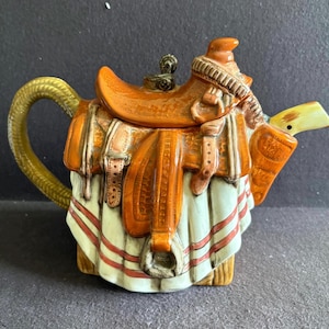 May include: A ceramic teapot shaped like a western saddle. The saddle is orange-brown with detailed straps and buckles. Below the saddle, a white cloth with horizontal red stripes drapes down. The handle is textured and yellow, and the spout is white.