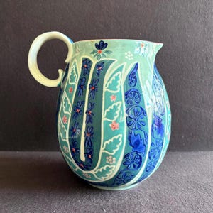 K&ouml;nnte beinhalten: Handbemalter Keramikkrug mit gebogenem Griff. Der Krug hat einen t&uuml;rkisfarbenen und teal-farbenen Hintergrund mit blauen und rosa Blumenmustern. Der Griff ist cremefarben.