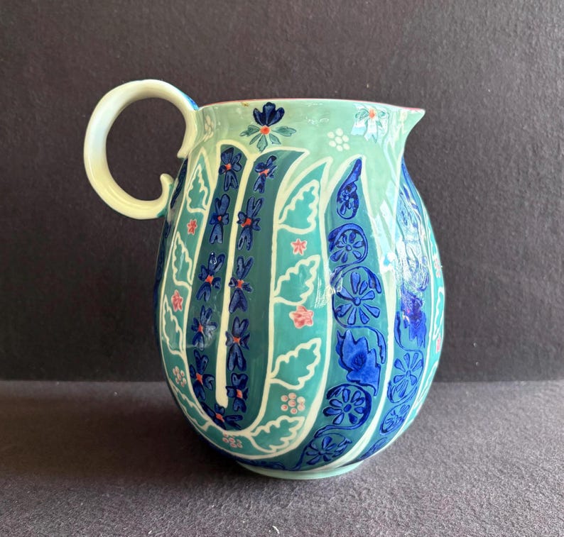 K&ouml;nnte beinhalten: Handbemalter Keramikkrug mit gebogenem Griff. Der Krug hat einen t&uuml;rkisfarbenen und teal-farbenen Hintergrund mit blauen und rosa Blumenmustern. Der Griff ist cremefarben.