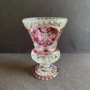 Puede incluir: Un jarrón de cristal transparente con la parte superior acampanada y una base redonda. El jarrón presenta un interior rojo rubí con un diseño floral blanco. La base y el borde tienen un acento dorado. El jarrón es probablemente vintage y es un objeto decorativo.