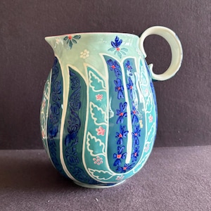 K&ouml;nnte beinhalten: Ein Keramikkrug mit rundem K&ouml;rper und gebogenem Henkel. Der Krug ist mit einem t&uuml;rkisfarbenen und blauen Blumendesign bemalt. Das Design umfasst stilisierte Blumen und Blattformen in Blau-, T&uuml;rkis- und Rosat&ouml;nen.