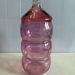 Puede incluir: Un tarro de cristal apilable de tres niveles en un tono rosa translúcido. Cada una de las tres secciones circulares presenta motivos florales grabados con hojas y flores. El tarro está rematado con una tapa de cristal extraíble a juego.