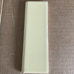 Op de afbeelding: Een rechthoekige, lichtgele keramische tegel met een glanzende afwerking. De tegel heeft een licht afgeronde rand aan één kant en een platte rand aan de andere kant. De tegel is effen van kleur en lijkt nieuw.