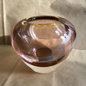 May include: A rounded glass vase featuring a translucent mauve upper section and a solid white base. A small rectangular label is affixed to the front, displaying the text "EVOLUTION BY WATERFORD" in clear, legible lettering on the glass surface.