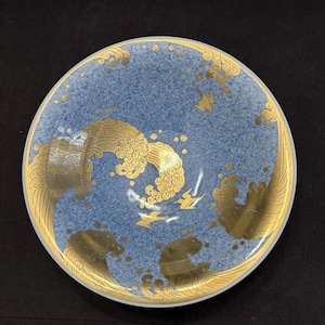 Puede incluir: Un plato redondo de cerámica con un fondo azul claro moteado y diseños dorados de olas y pájaros. El borde y los detalles de las olas están delineados en dorado. El plato es una pieza decorativa.