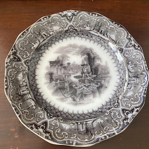 May include: A vintage plate featuring a black and white landscape design. The plate has a scalloped edge and an ornate border with architectural and floral patterns. The plate's design suggests it is an antique piece.