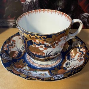 Puede incluir: Un juego de taza y platillo detallado. La taza y el platillo presentan un fondo azul oscuro con detalles dorados y diseños intrincados. Los diseños incluyen elementos florales y arquitectónicos naranjas y blancos. La taza tiene un interior blanco y un borde festoneado.