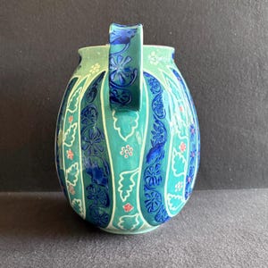 K&ouml;nnte beinhalten: Ein Keramikkrug mit einem t&uuml;rkisblauen Blumenmuster. Der Krug hat einen abgerundeten K&ouml;rper mit einem Henkel und ist mit vertikalen Streifen aus blauen Blumenmustern und kleinen rosa Akzenten verziert. Der Hintergrund ist dunkel.