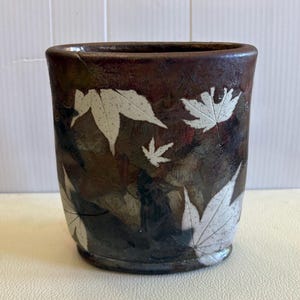 Peut inclure: Un gobelet en céramique marron avec une finition brillante. Le gobelet présente un motif de feuilles d'érable blanches. Les feuilles sont dispersées sur la surface du gobelet. Le gobelet a une forme légèrement rectangulaire.