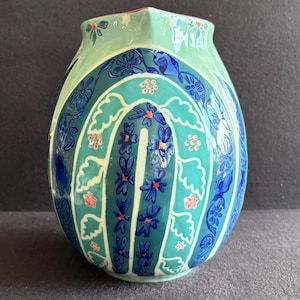 K&ouml;nnte beinhalten: Eine dekorative Keramikvase mit einer einzigartigen Form und einem farbenfrohen Blumendesign. Die Vase hat eine hellgr&uuml;ne Oberseite, einen t&uuml;rkisfarbenen Mittelteil und einen dunkelblauen Sockel. Das Design umfasst blaue Blumen und rosa Akzente.