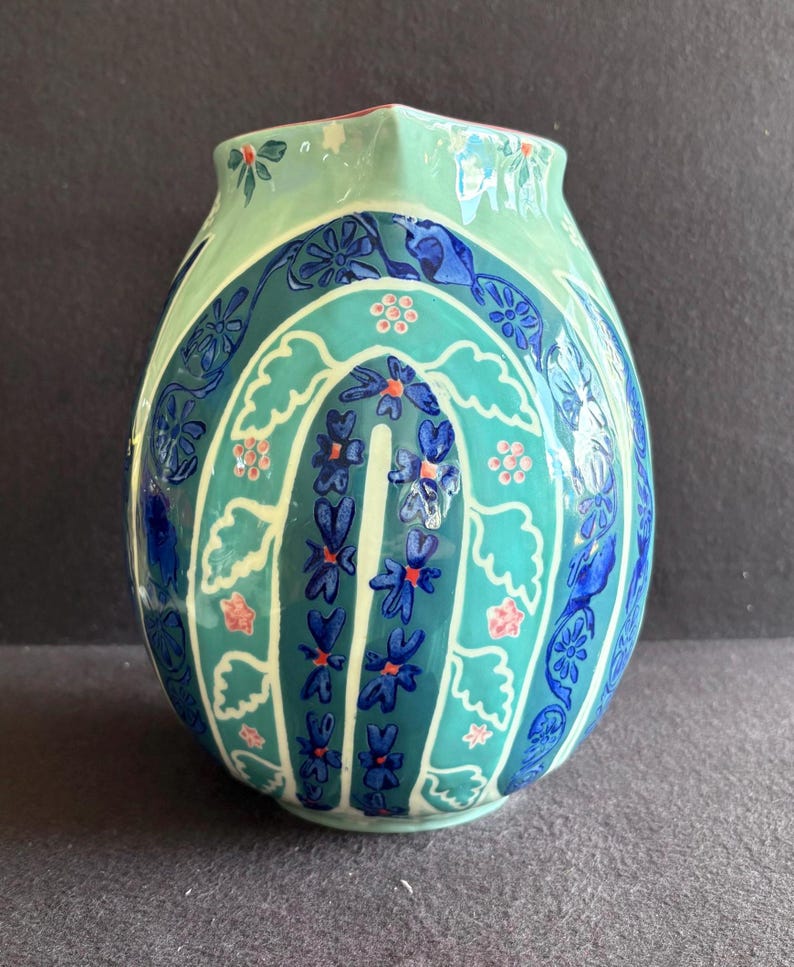 K&ouml;nnte beinhalten: Eine dekorative Keramikvase mit einer einzigartigen Form und einem farbenfrohen Blumendesign. Die Vase hat eine hellgr&uuml;ne Oberseite, einen t&uuml;rkisfarbenen Mittelteil und einen dunkelblauen Sockel. Das Design umfasst blaue Blumen und rosa Akzente.