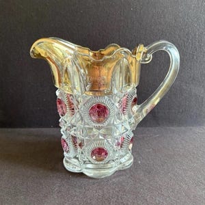Op de afbeelding: Een heldere glazen creamer met een gouden rand en handvat. De creamer heeft een diamantgeslepen patroon met rode cirkelvormige accenten. Het handvat is gebogen en de basis is geschulpt. Een decoratief stuk.