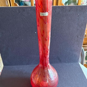 May include: A tall, slender red glass vase with a bulbous base and a long, tapering neck. The vase has a swirling pattern throughout and a label that reads "EVOLUTION BY WATERFORD".