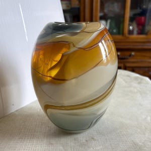 May include: A decorative glass vase with a rounded shape. The vase features a blend of colors, including amber, cream, gray, and brown, with swirling patterns. The vase has a glossy finish and is suitable for home decor.