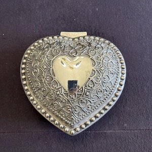 May include: Silver-toned heart-shaped trinket box with ornate filigree detailing. The box features a central heart-shaped accent in a lighter silver tone. The edges are lined with small, raised beads. The box is closed with a small clasp.